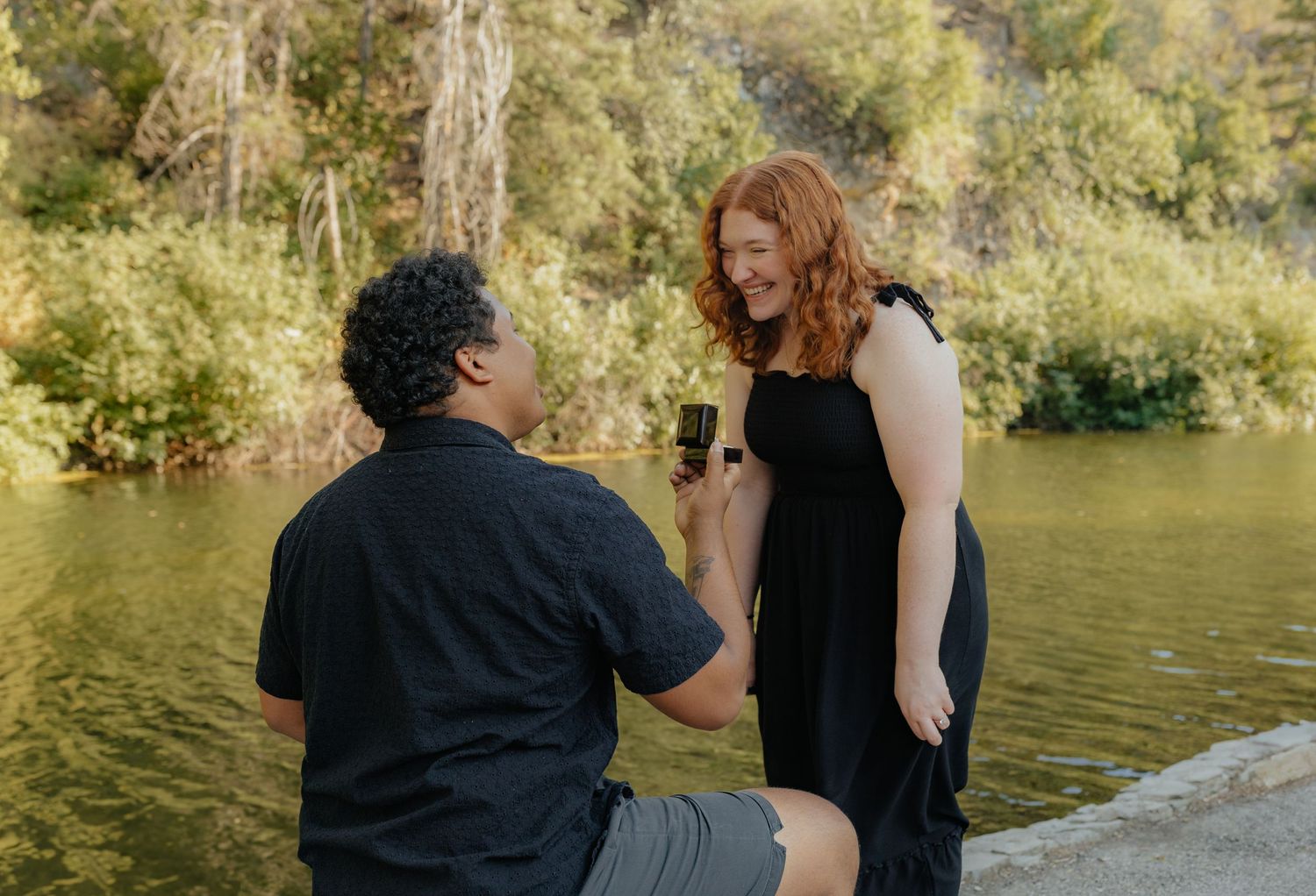 Man proposes on one knee to redhead woman in black dress near scenic lakeside setting.
