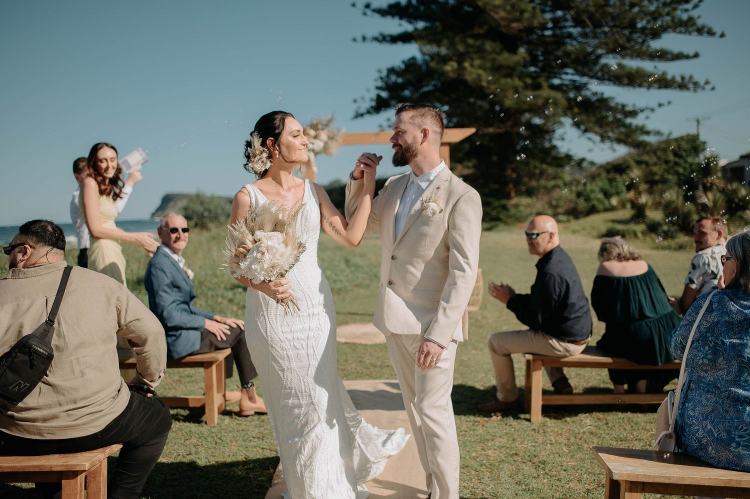 Couple sharing celebratory toast outdoors with wedding guests seated on benches in background.