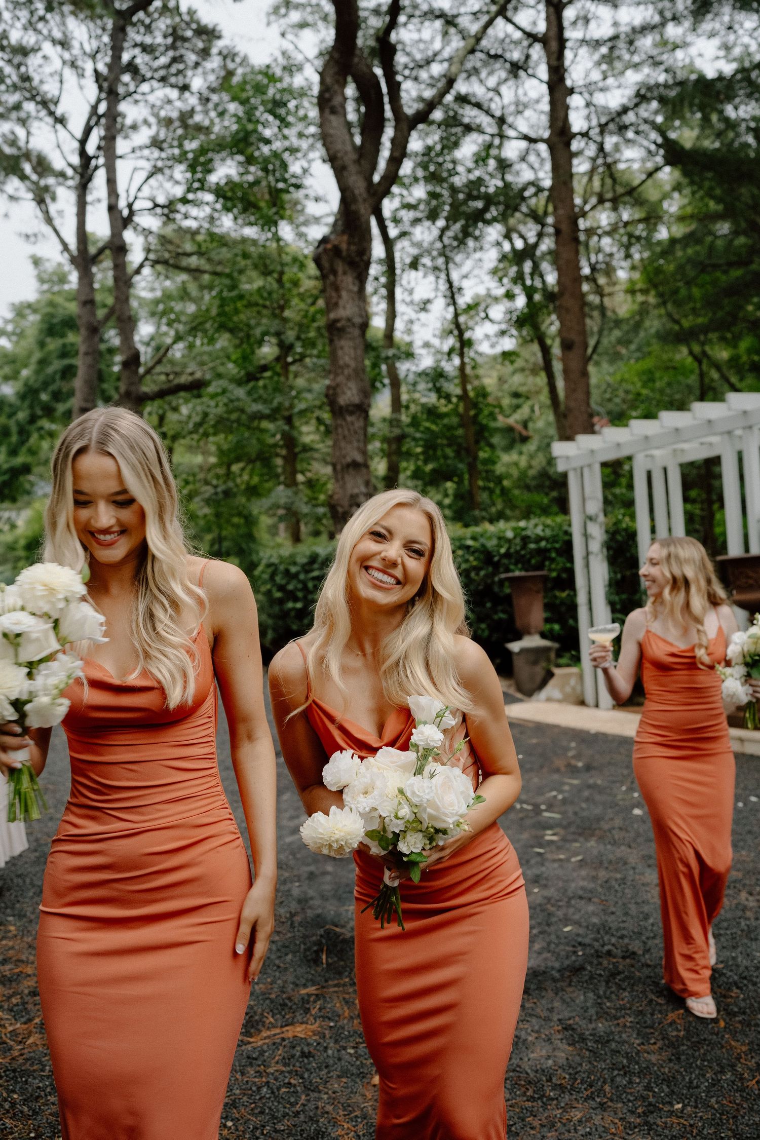 Bridesmaids in fitted rust-colored dresses laughing together at an outdoor wedding venue with trees.