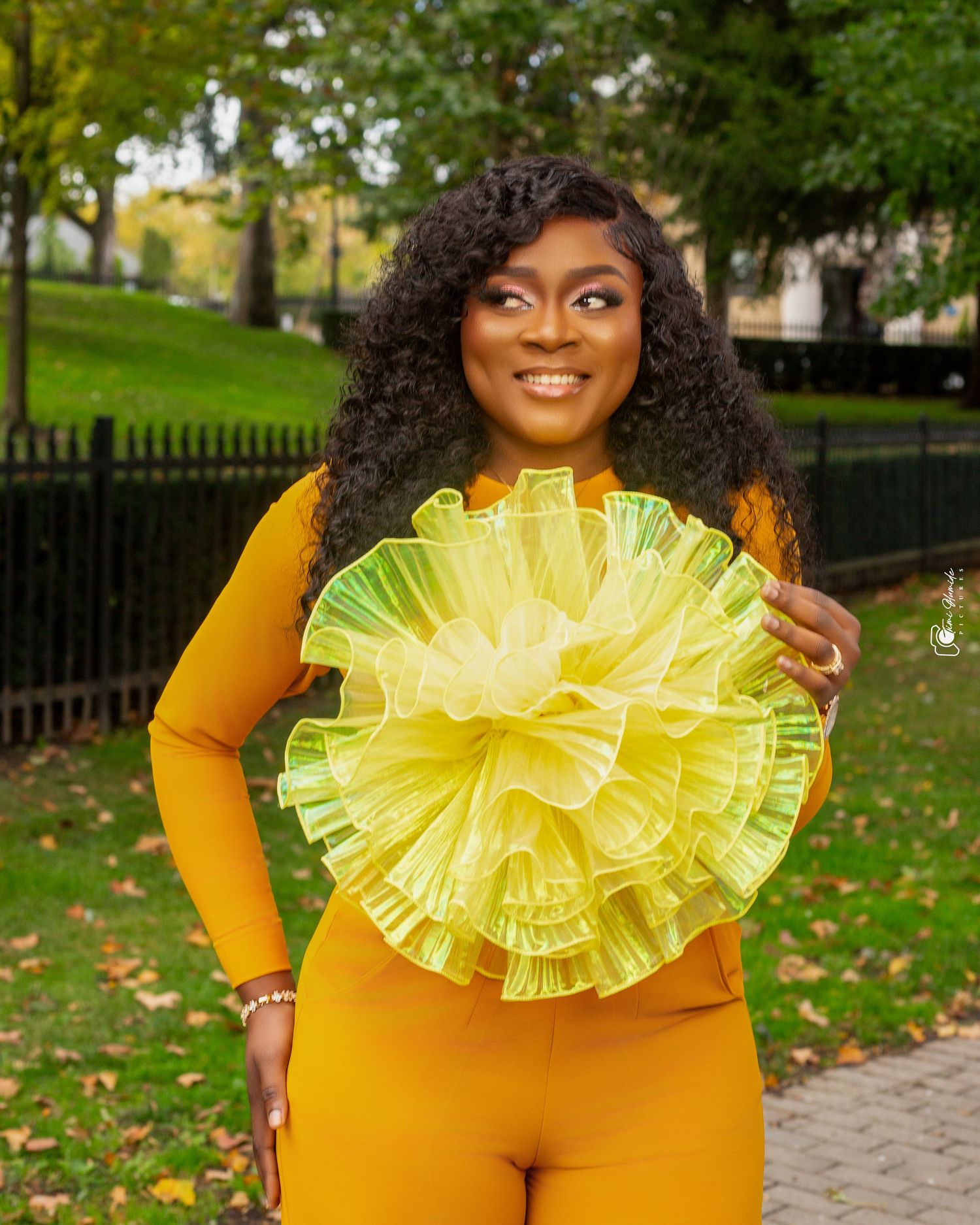 Portrait of someone in bright yellow outfit holding large decorative yellow pom-pom outdoors.