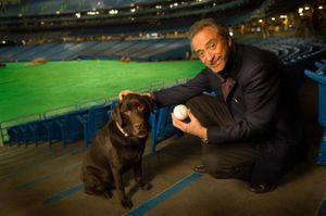 A person in a suit kneeling next to a chocolate Labrador dog in a stadium.