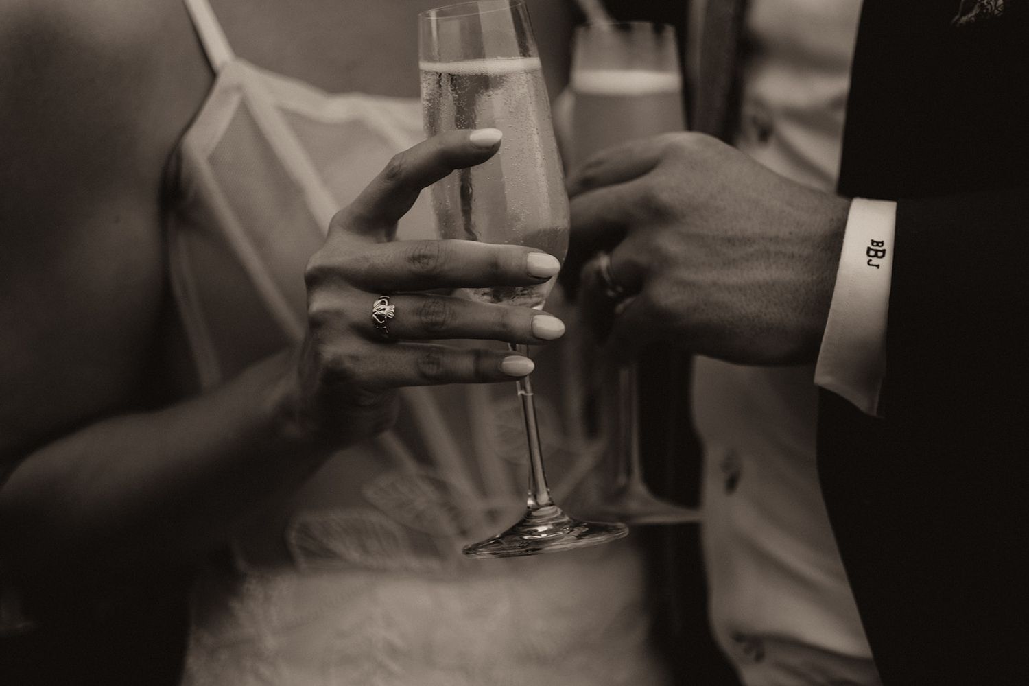 Close-up of bride and groom holding champagne flutes, showing wedding ring, black and white photo.