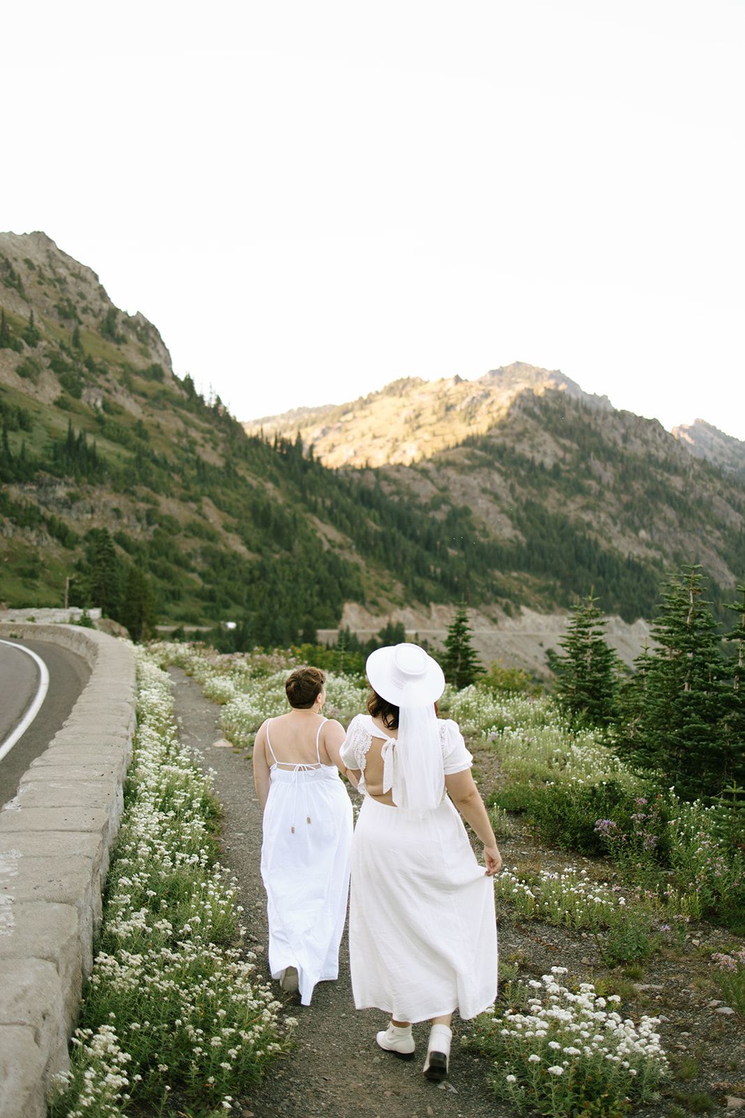 Two brides in white dresses walk together along mountain road surrounded by wildflowers at sunset.