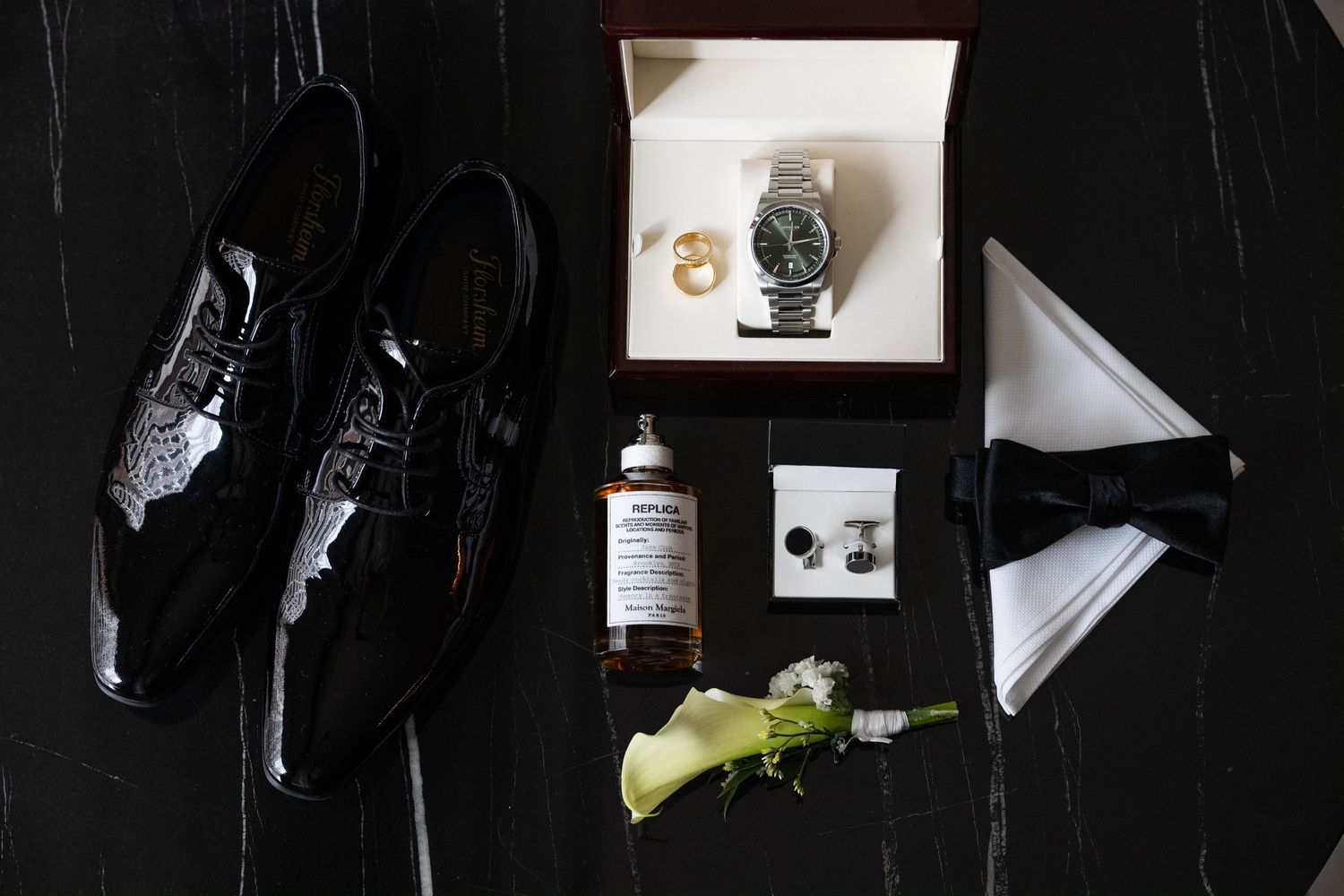 Wedding accessories including dress shoes, watch, boutonniere and whiskey bottle arranged on dark surface.