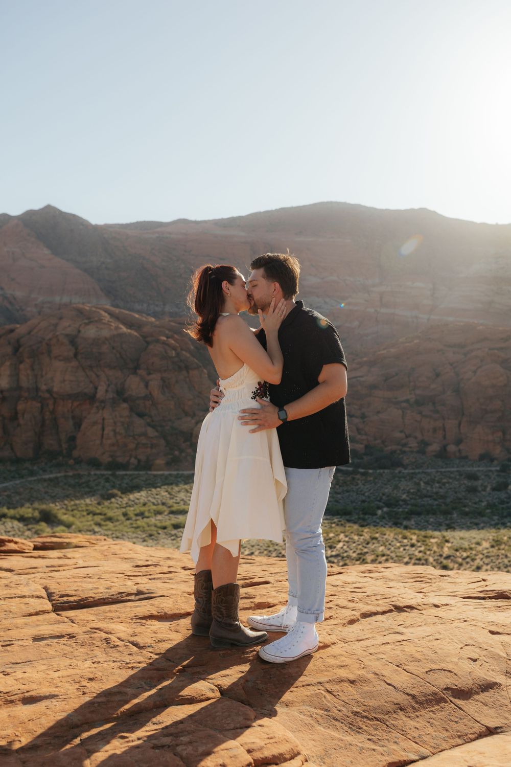 Bride in white dress and cowboy boots kisses groom on red rock overlook with mountain backdrop.