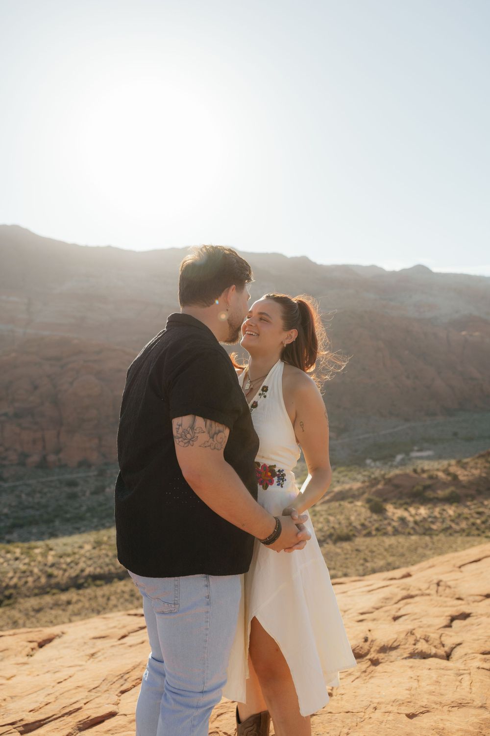 Bride in white halter dress smiles at tattooed groom on sunlit red rock desert landscape.