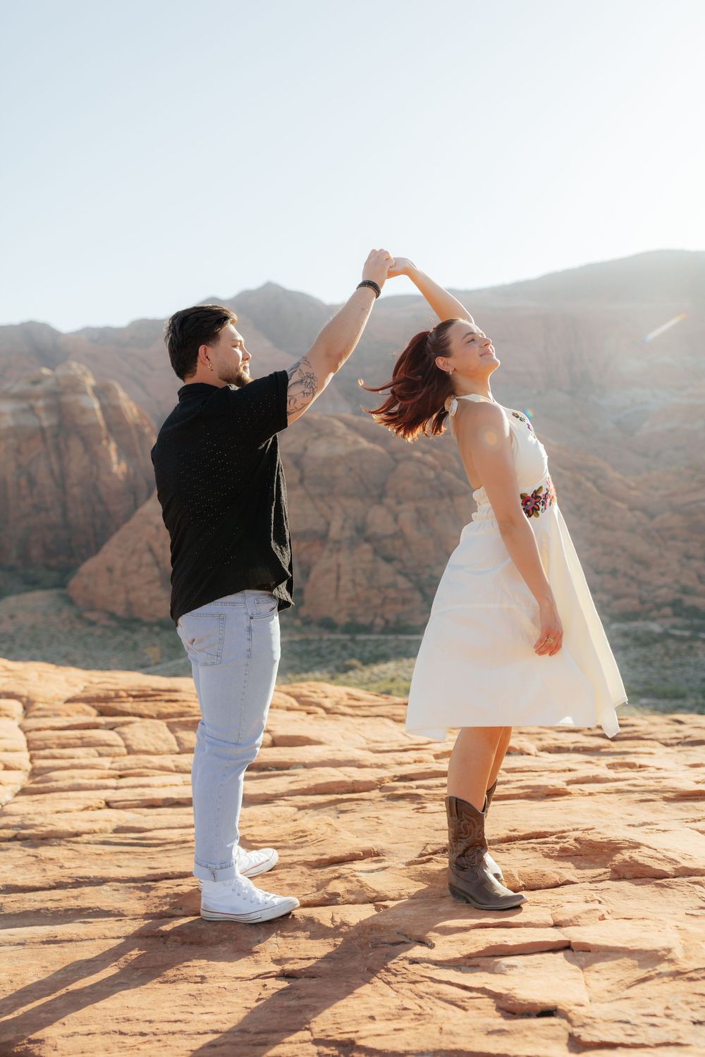 Man spinning woman in a white dress and cowboy boots on red rock desert terrain at sunset.