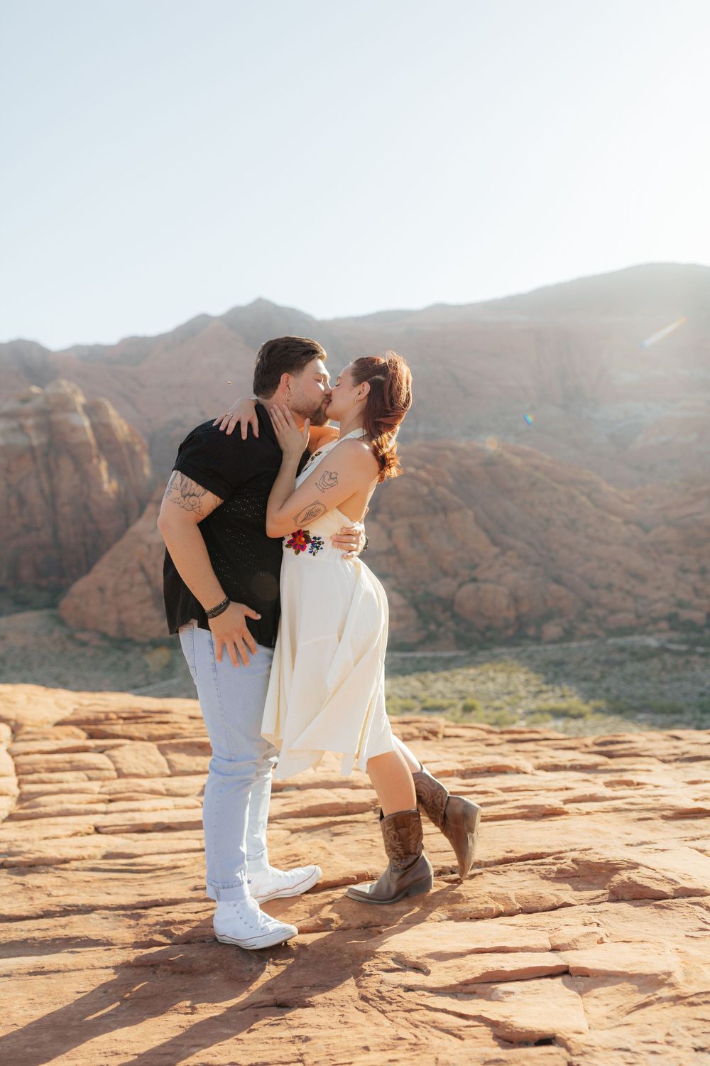 Couple sharing a romantic kiss on red rock formations with stunning desert canyon landscape backdrop.