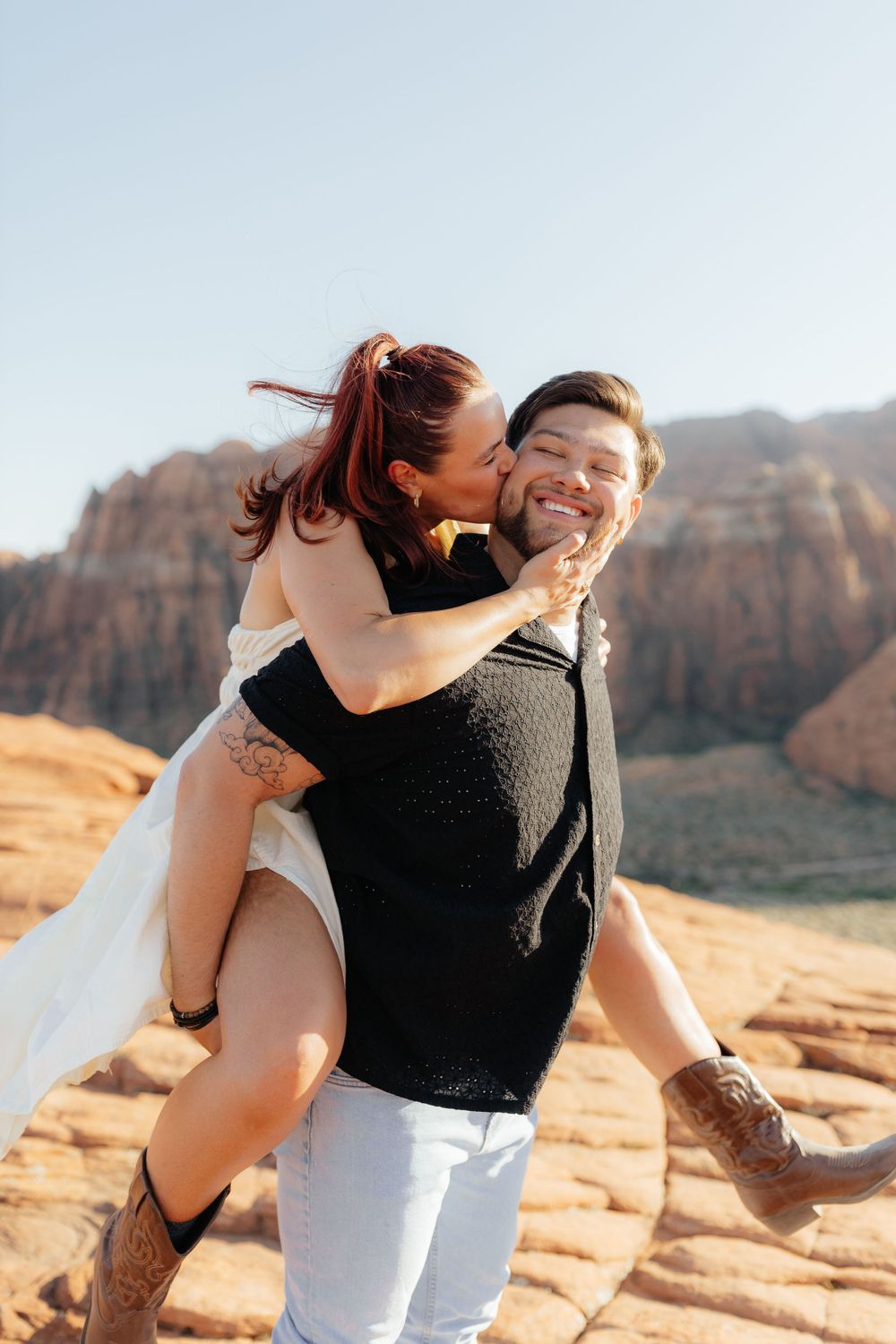 Groom smiles as bride kisses his cheek while being carried piggyback on red rock desert terrain.