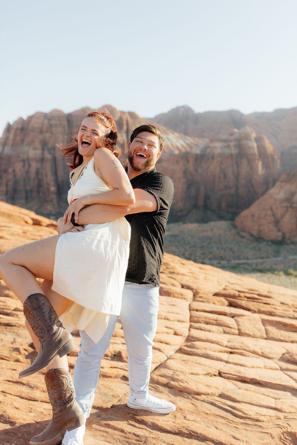 Joyful couple laughing as man lifts woman in white dress and cowboy boots at red rock canyon.