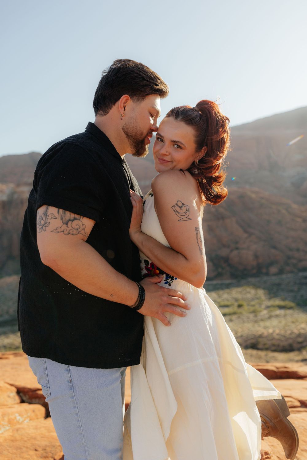 Couple embracing on red rock desert landscape, woman in white dress with floral embroidery smiling at camera.
