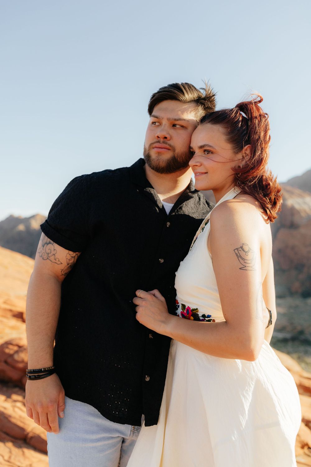 Man in black shirt and woman in white halter dress pose together against red rock desert scenery.