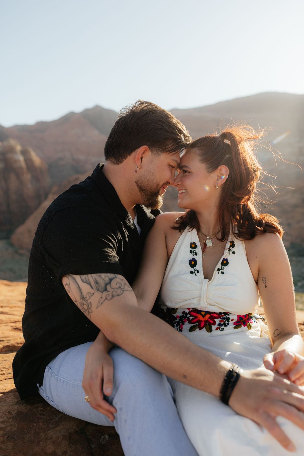 Couple sitting on red rock, leaning foreheads together and smiling in a sunny desert canyon setting.