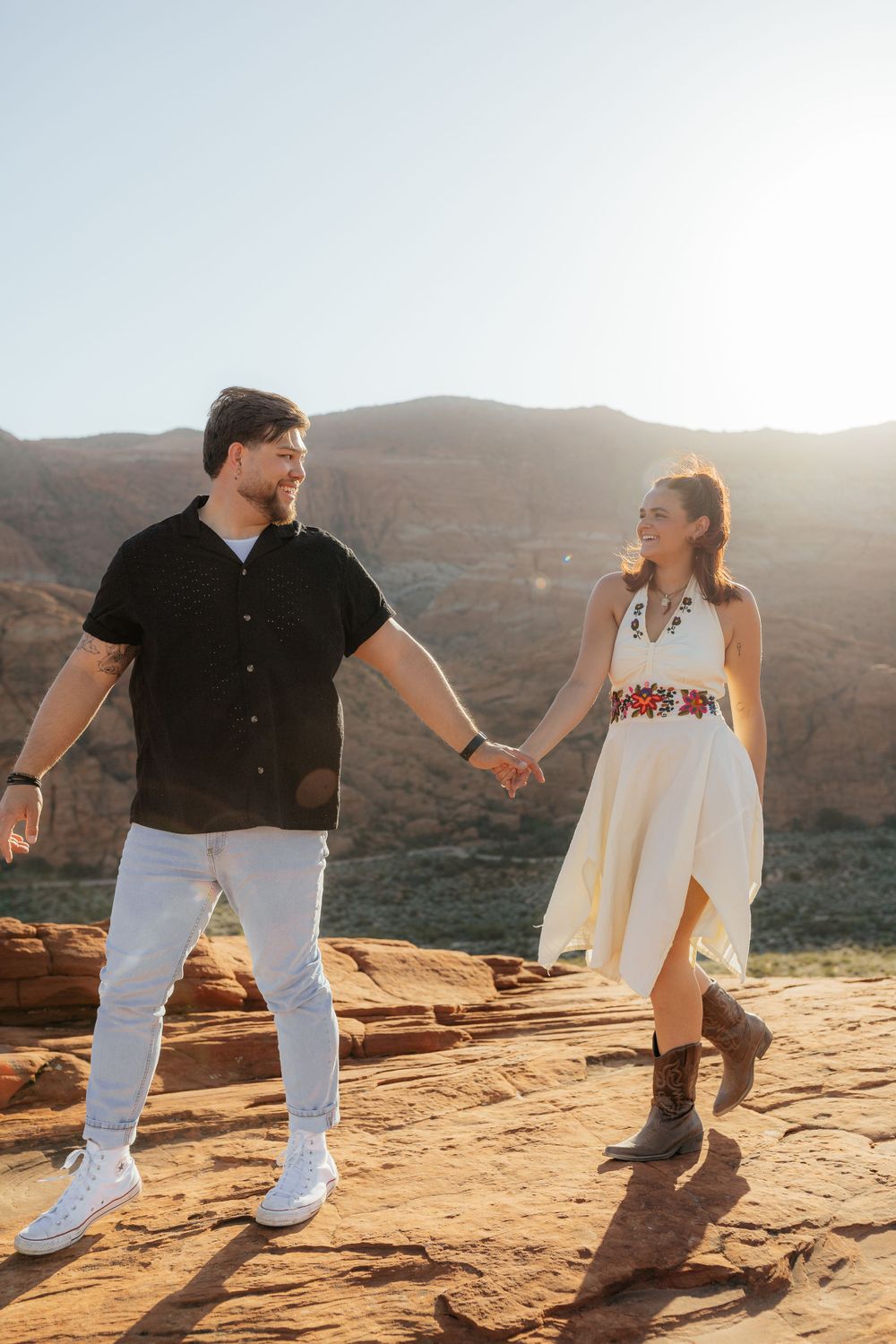 Couple holding hands and walking on red sandstone with sunlit canyon mountains behind them.