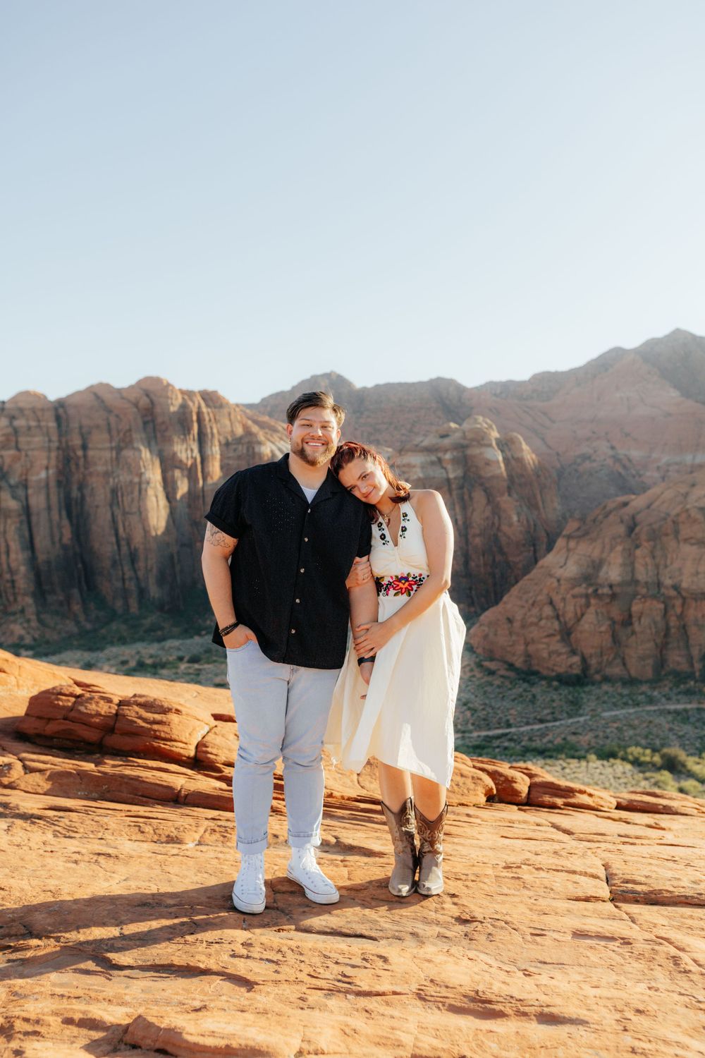 Smiling couple posing together on red sandstone with dramatic red rock canyon mountains behind them.