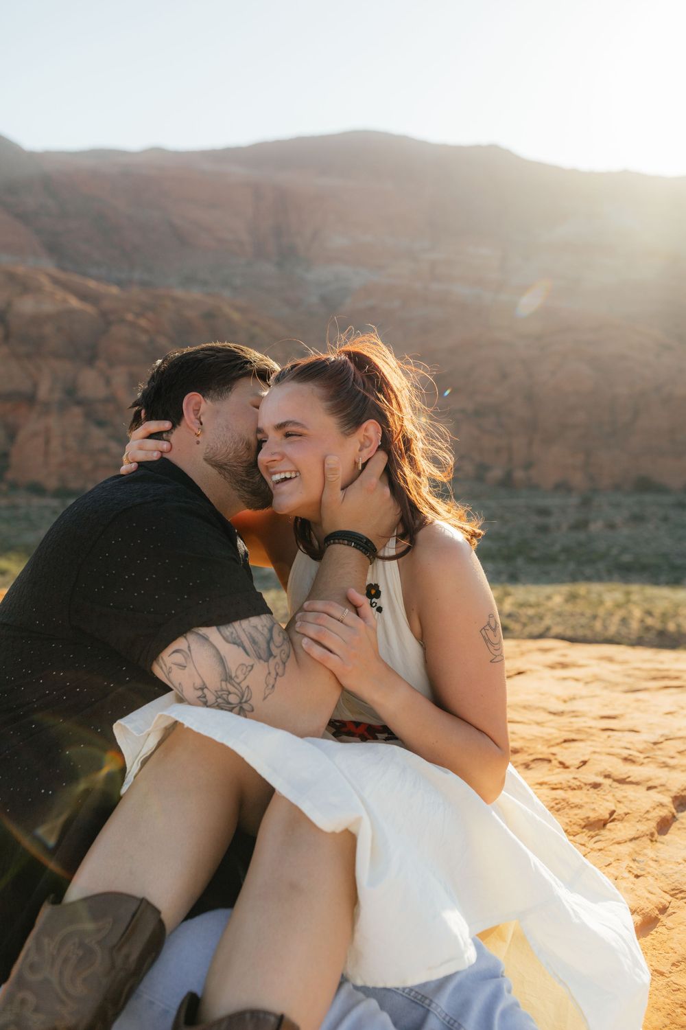 Man whispering to laughing woman while sitting together on sandy desert ground at golden hour.