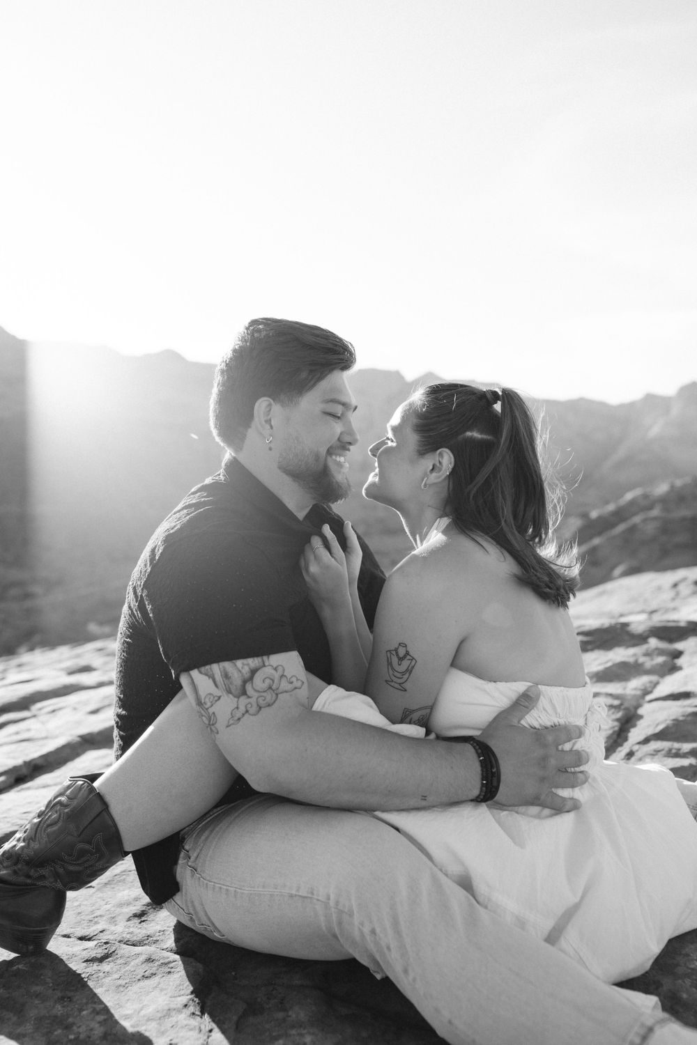 Black and white photo of couple sitting on rocks, smiling face to face with bright backlight behind them.