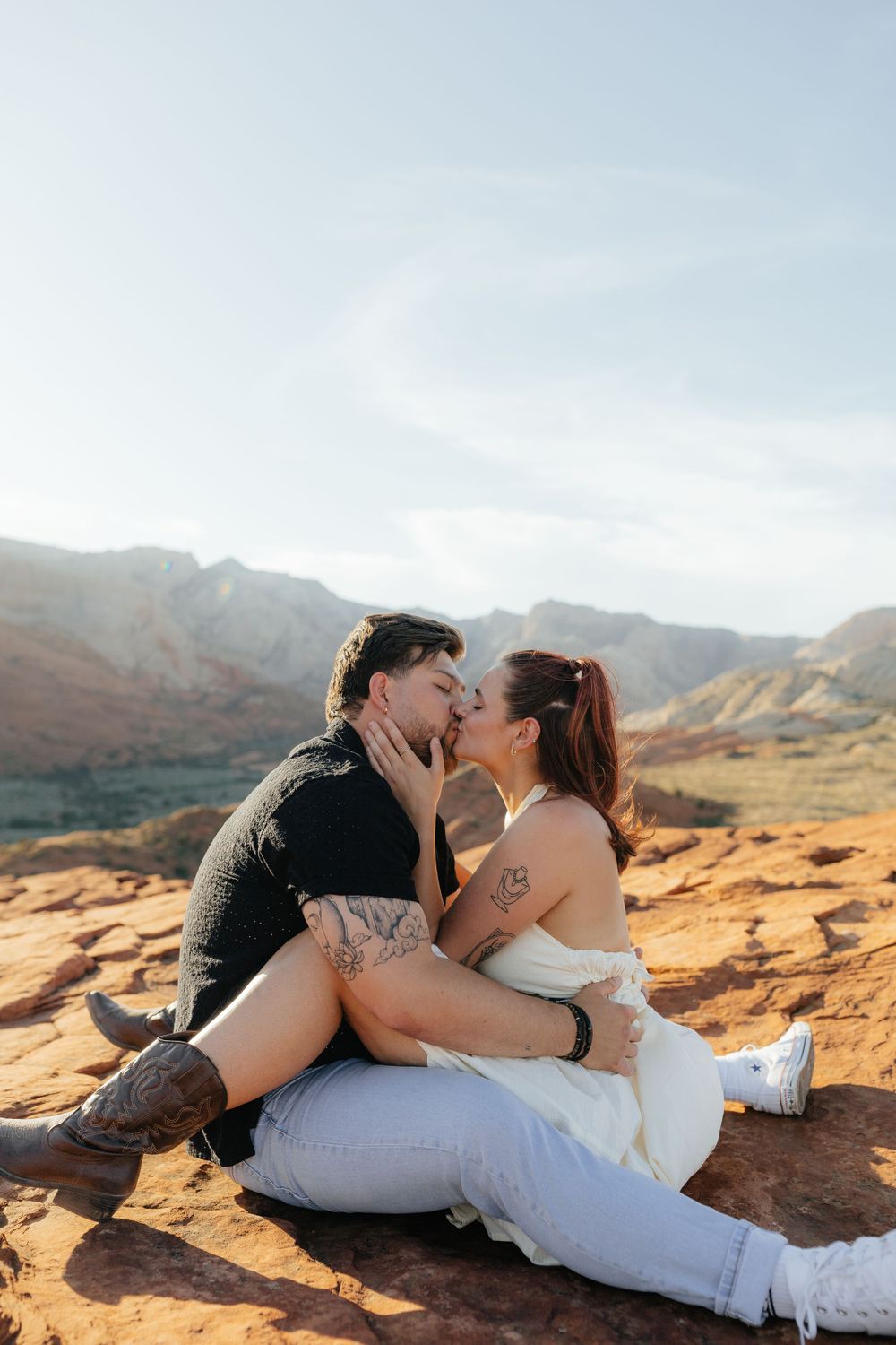 Couple sharing a passionate kiss while sitting on red sandstone with mountain landscape behind them.