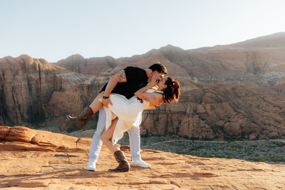 Man dipping woman in a romantic kiss on red rock overlook with sunlit canyon mountains in background.