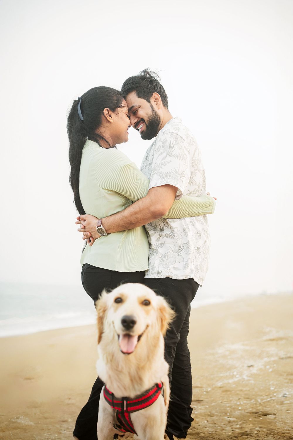 A happy couple embraces on the beach with their smiling golden retriever.
