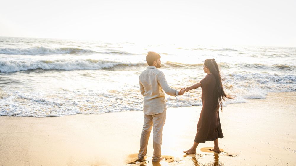 A couple holds hands while walking along a beach at sunset with waves crashing behind them.