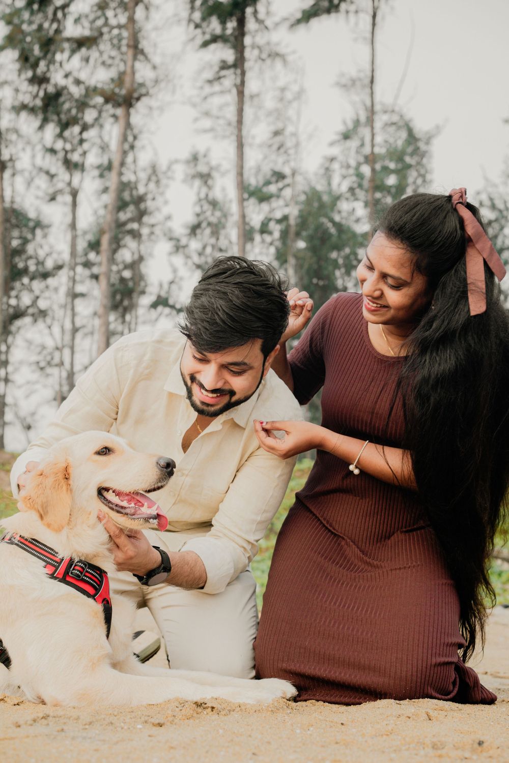 A couple sits on grass with their happy Golden Retriever during an outdoor photoshoot in a forest setting.