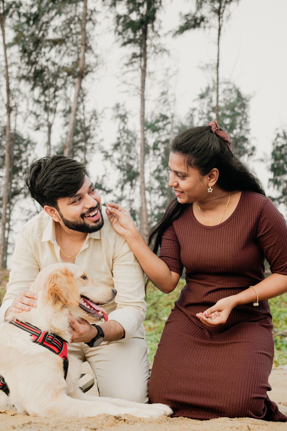 A joyful couple shares a playful moment with their Golden Retriever dog against a backdrop of tall trees.