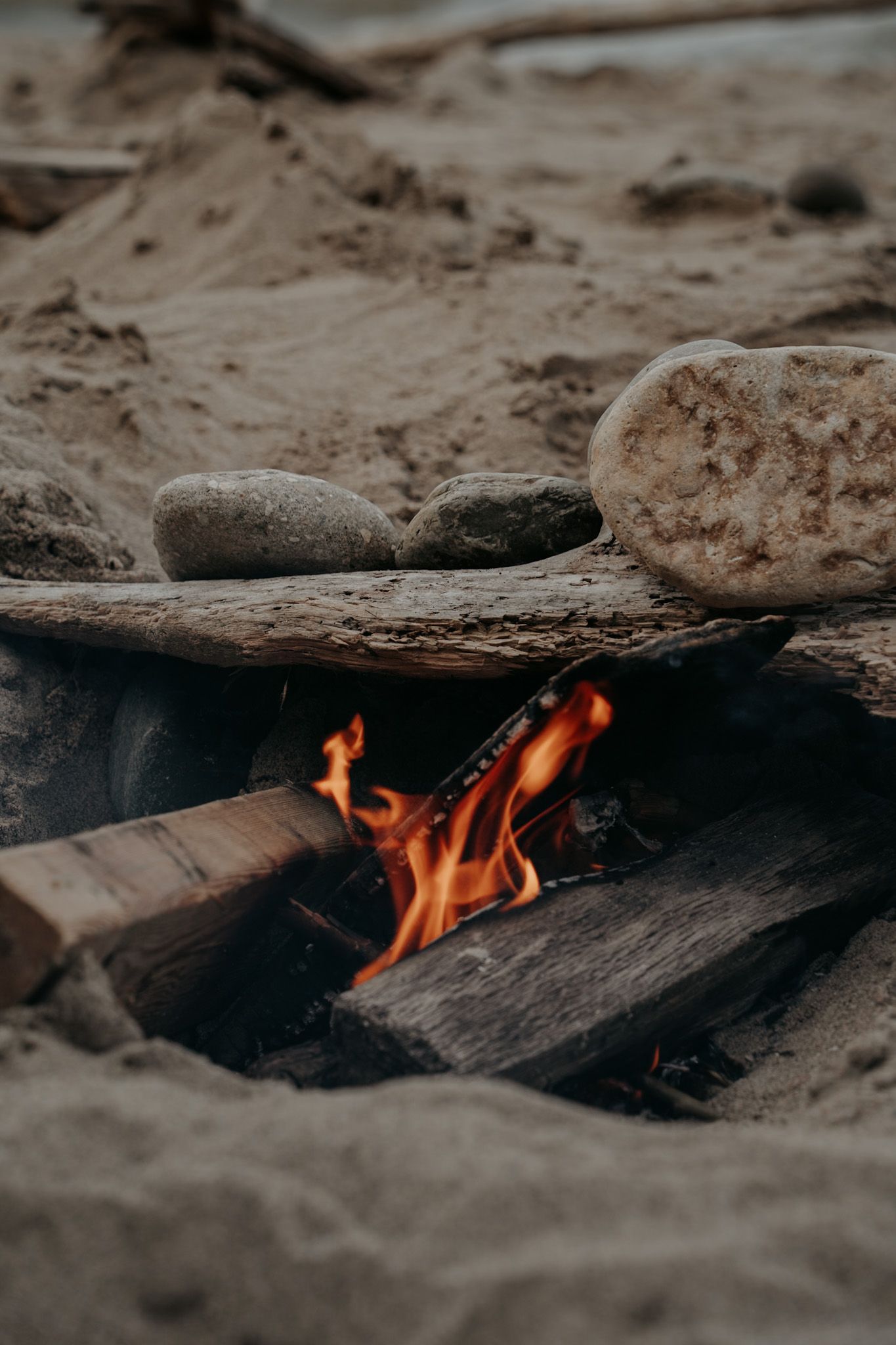 A close-up of a beach campfire with flames licking wooden logs surrounded by sand and stones.