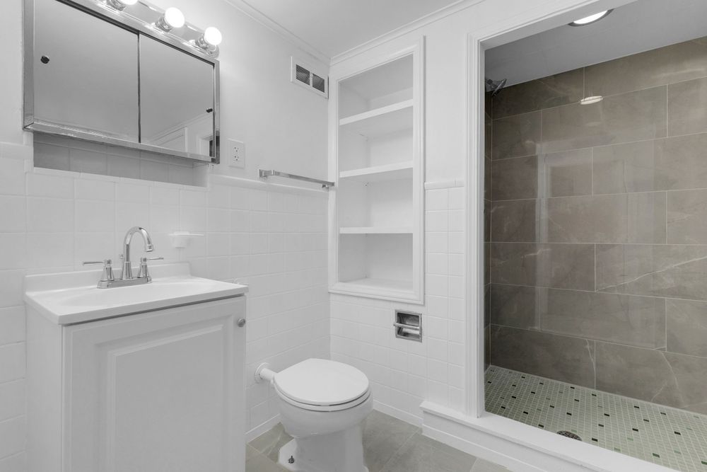 Modern white bathroom with vanity, medicine cabinet, toilet, built-in shelves, and tiled walk-in shower.