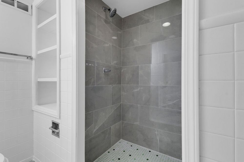 Spacious walk-in shower with large gray tiles, mosaic floor, built-in shelves, and recessed lighting.