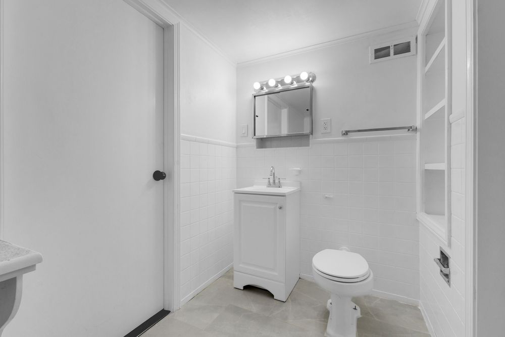 Clean white bathroom with vanity, mirror cabinet, toilet, and built-in shelving on light tile floors.