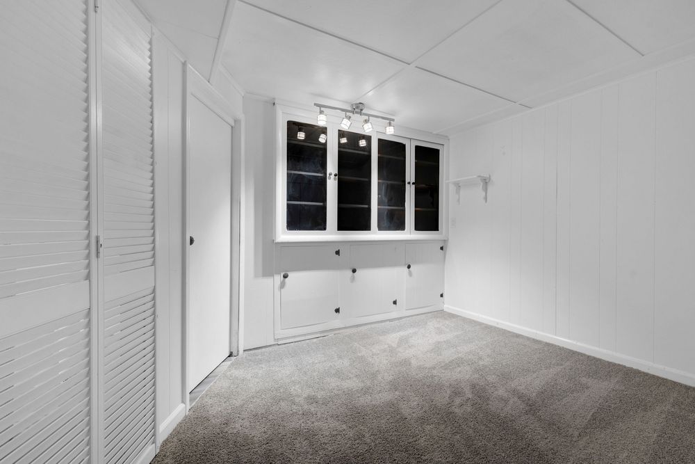 Empty room with gray carpet, white louvered closet doors, built-in white cabinets, and a window with black trim.