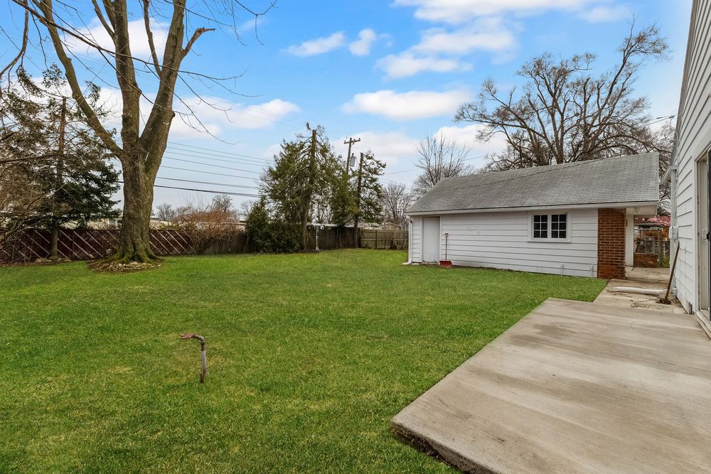 Spacious backyard with lush green lawn, detached garage, wooden fence, and mature trees under a blue sky.