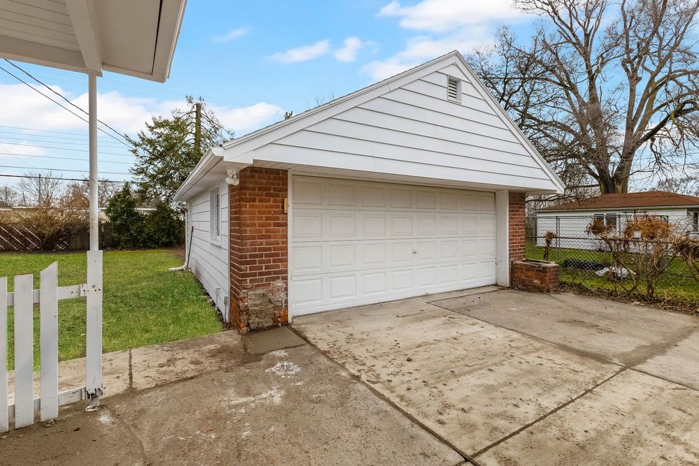 Detached brick and white garage with concrete driveway beside white picket fence in suburban yard.