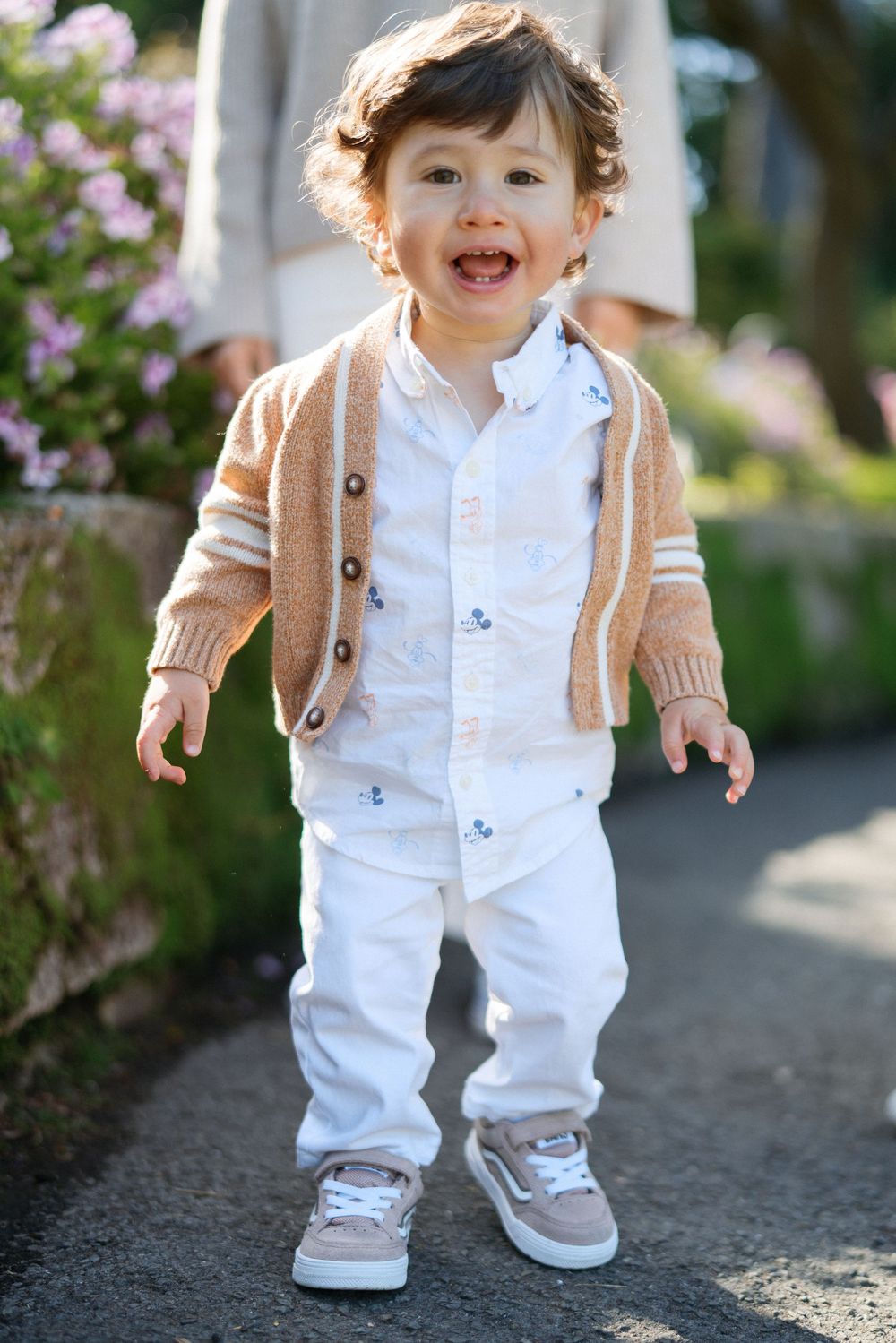Smiling toddler in beige cardigan and white pants standing on garden path in natural light.