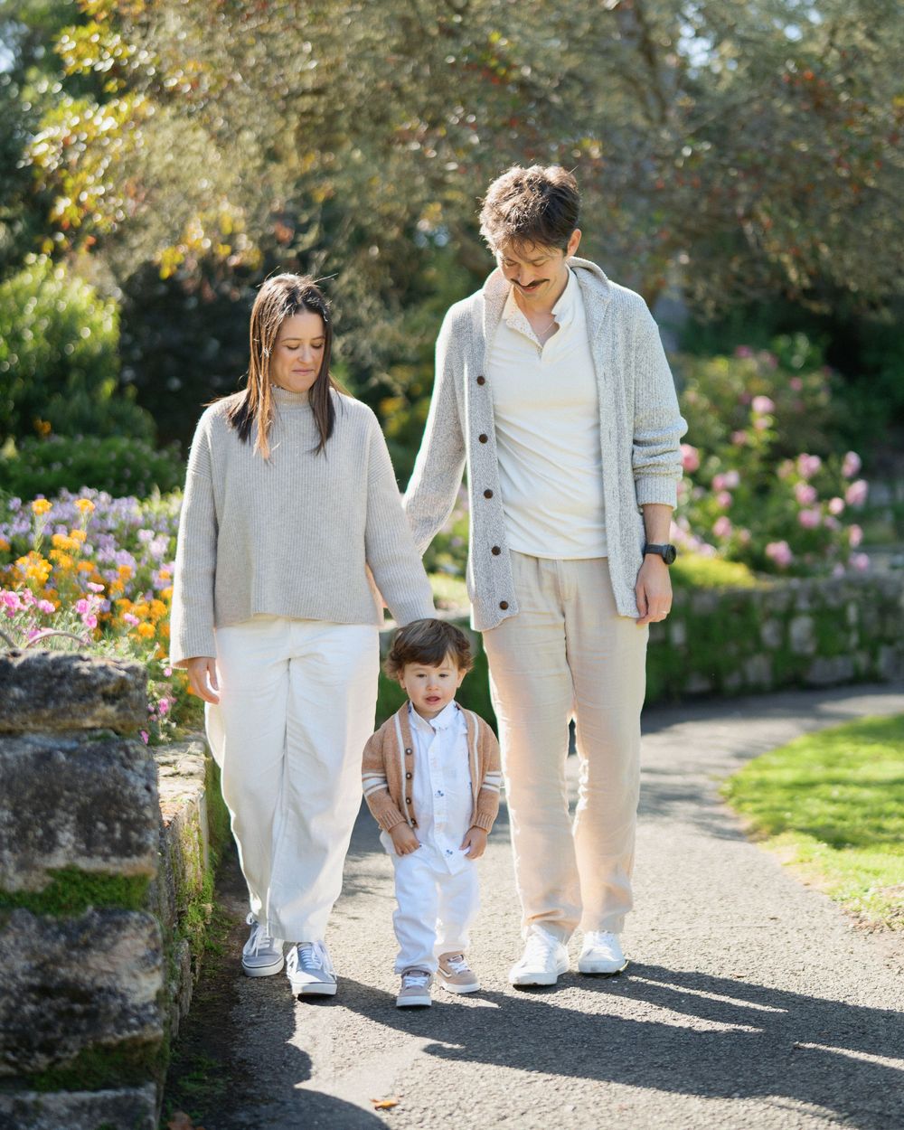 Family of three walking through a blooming garden, parents holding toddler's hands, dressed in neutral tones.