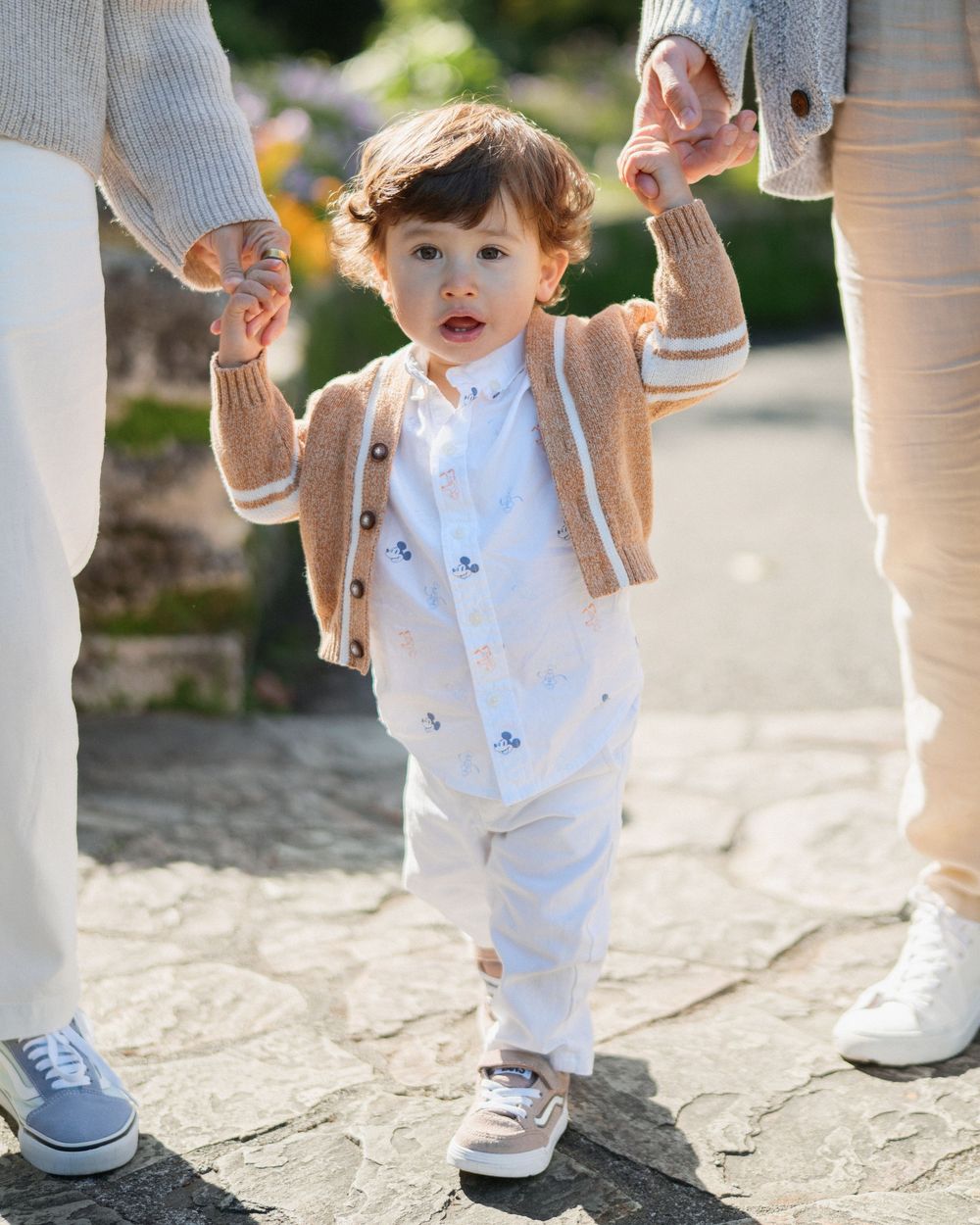 Adorable toddler in beige cardigan and white outfit walking while holding parents hands outdoors.
