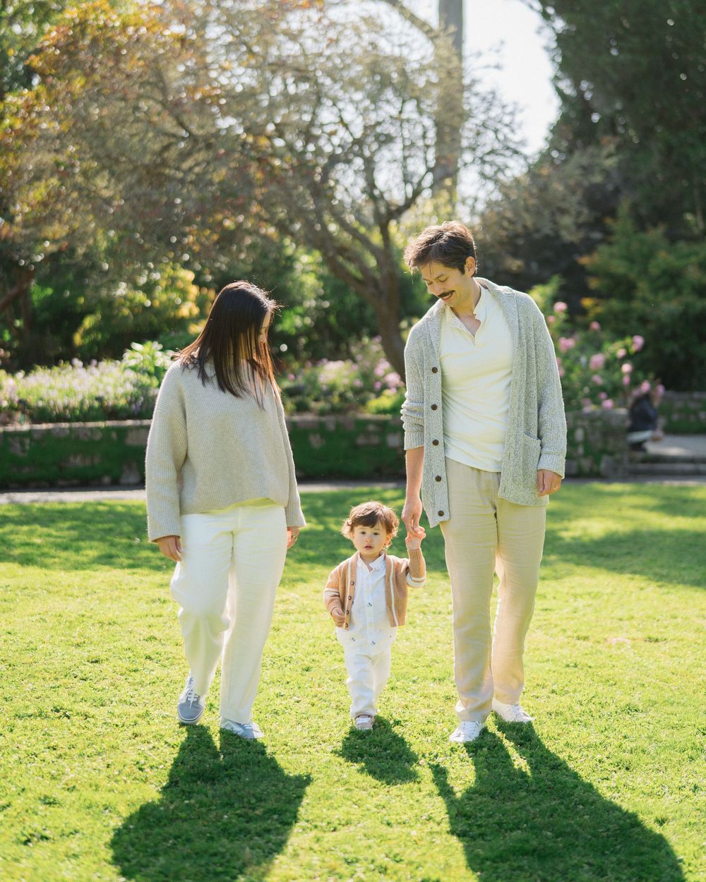 Family of three enjoying a sunny day in a lush green park, parents holding toddler's hands.