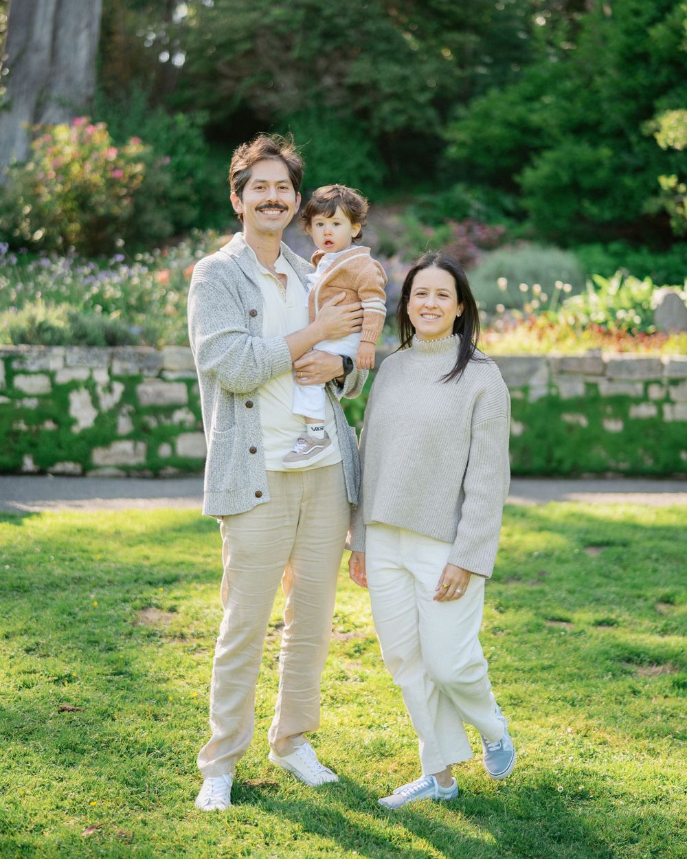 Happy family of three in neutral outfits pose together in a sunlit garden, man cradling a young toddler.