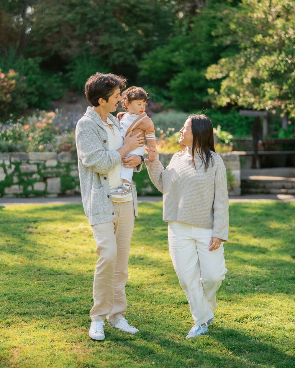 Family of three posing on green lawn in a garden, parents in neutral tones holding toddler.