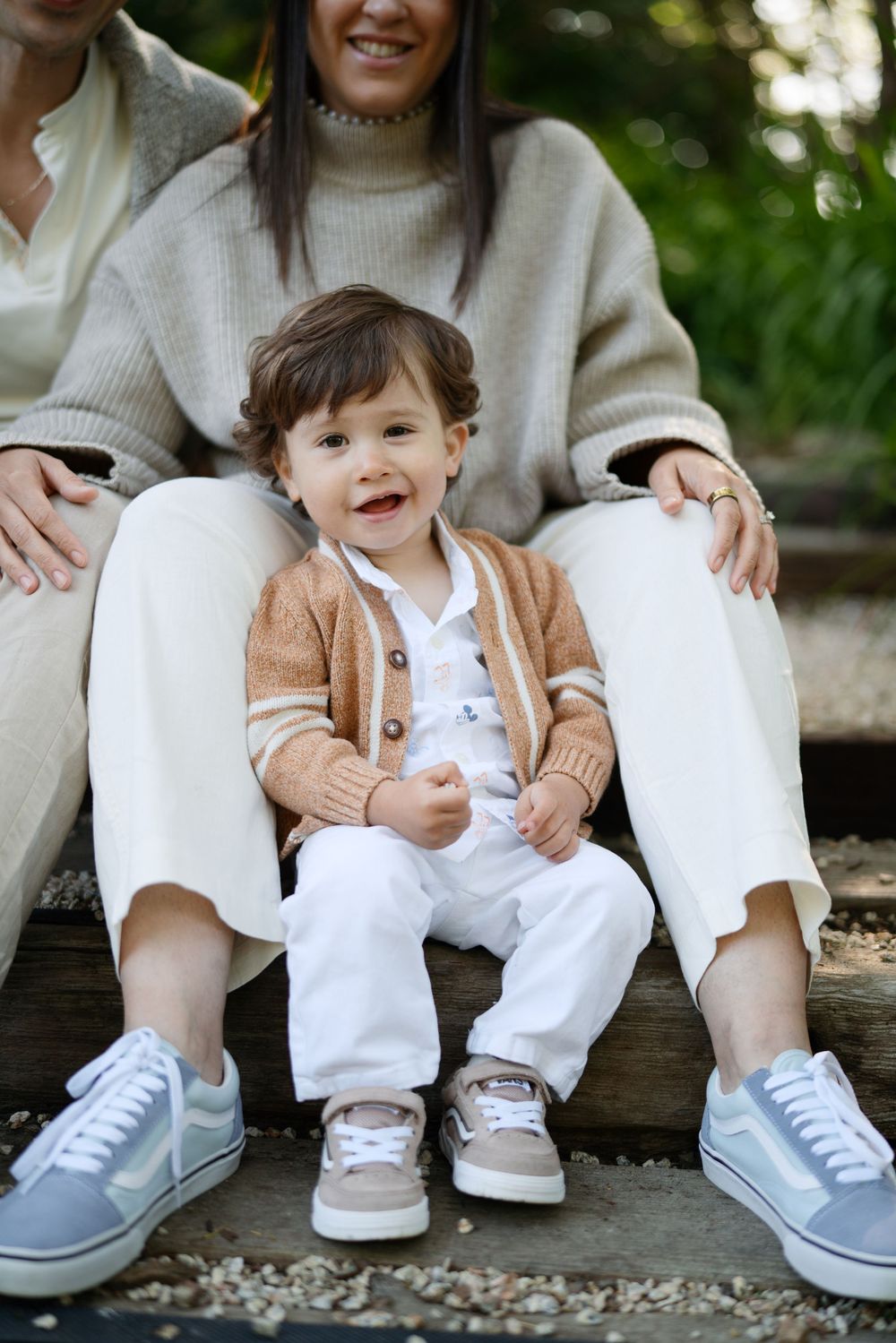 Smiling toddler boy in brown cardigan sits between two women in white outfits on wooden steps outdoors.