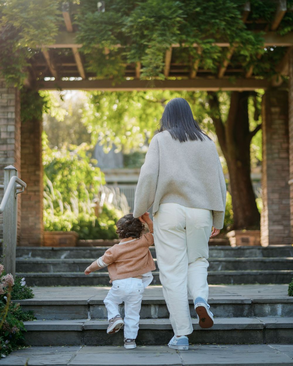 Woman and toddler walking hand in hand up stone steps under a pergola in a garden setting.