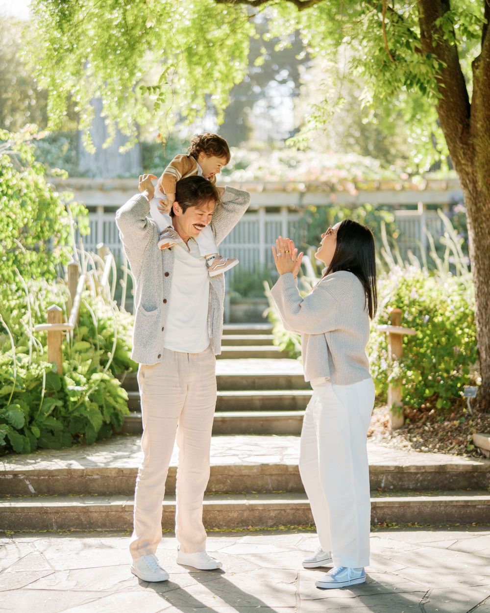 Parents lifting toddler overhead outdoors, dressed in coordinated white and gray outfits in garden.