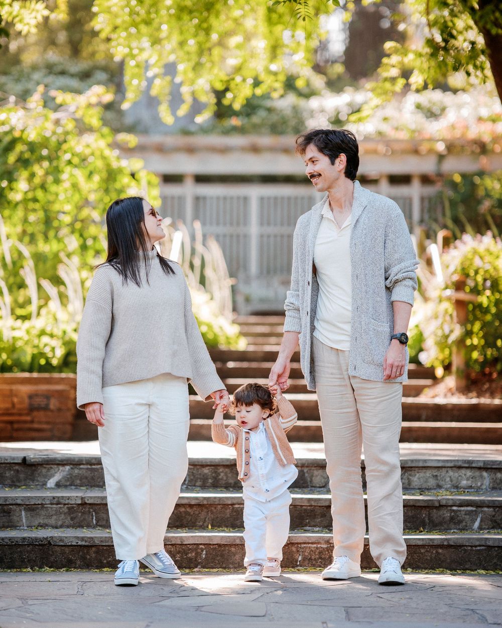 A young family with a toddler holds hands on garden steps, dressed in neutral tones, smiling warmly outdoors.