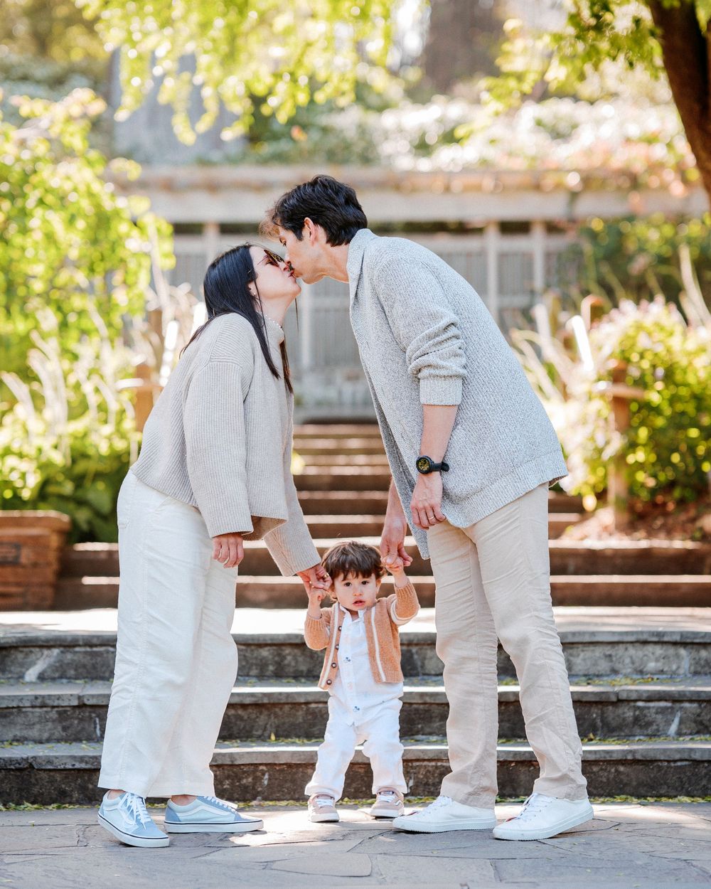 A young family in matching beige outfits shares a kiss over their toddler in a sunlit garden.