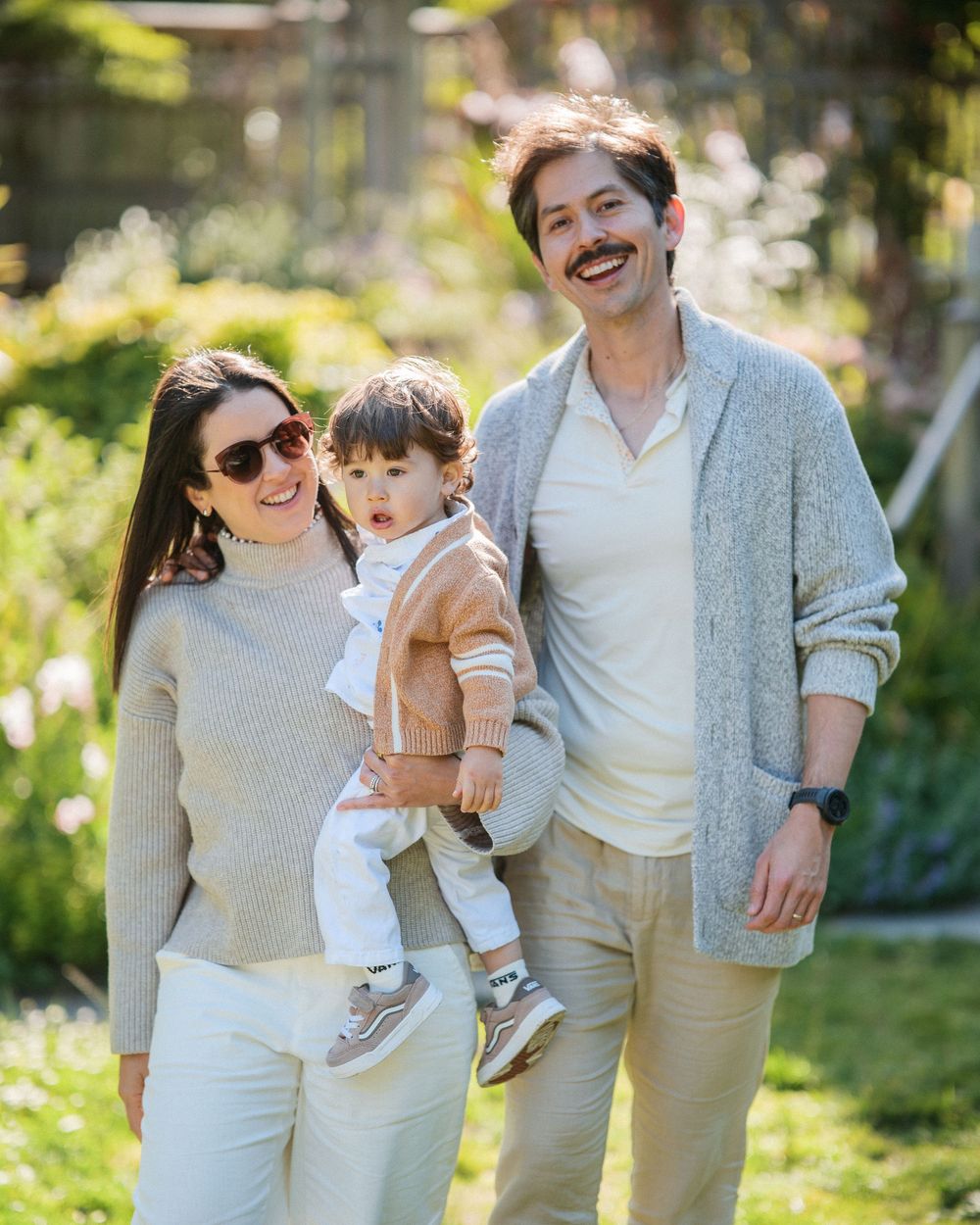 Happy family of three posing outdoors in neutral tones, parents holding toddler in sunlit garden.