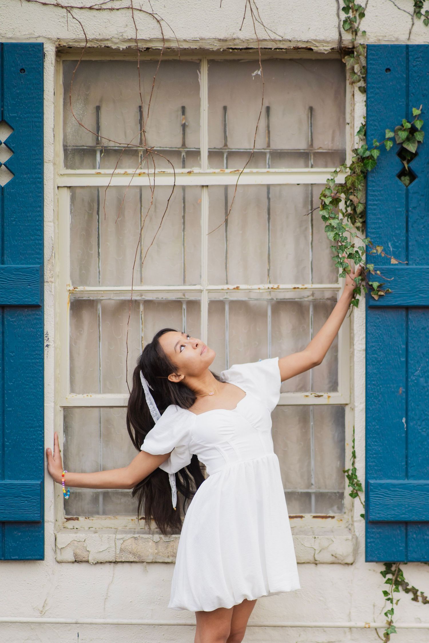 Young woman in white dress poses gracefully beside a rustic window with blue shutters and ivy vines.