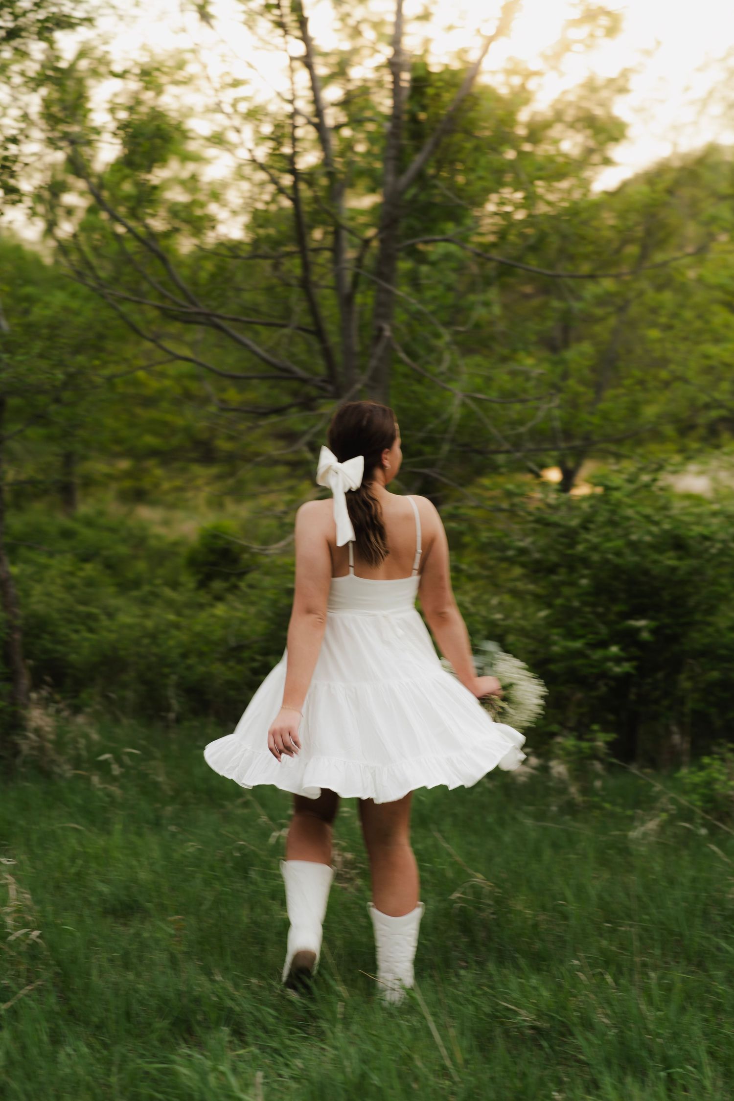 Young woman in white mini dress and white boots walks through a lush green meadow from behind.
