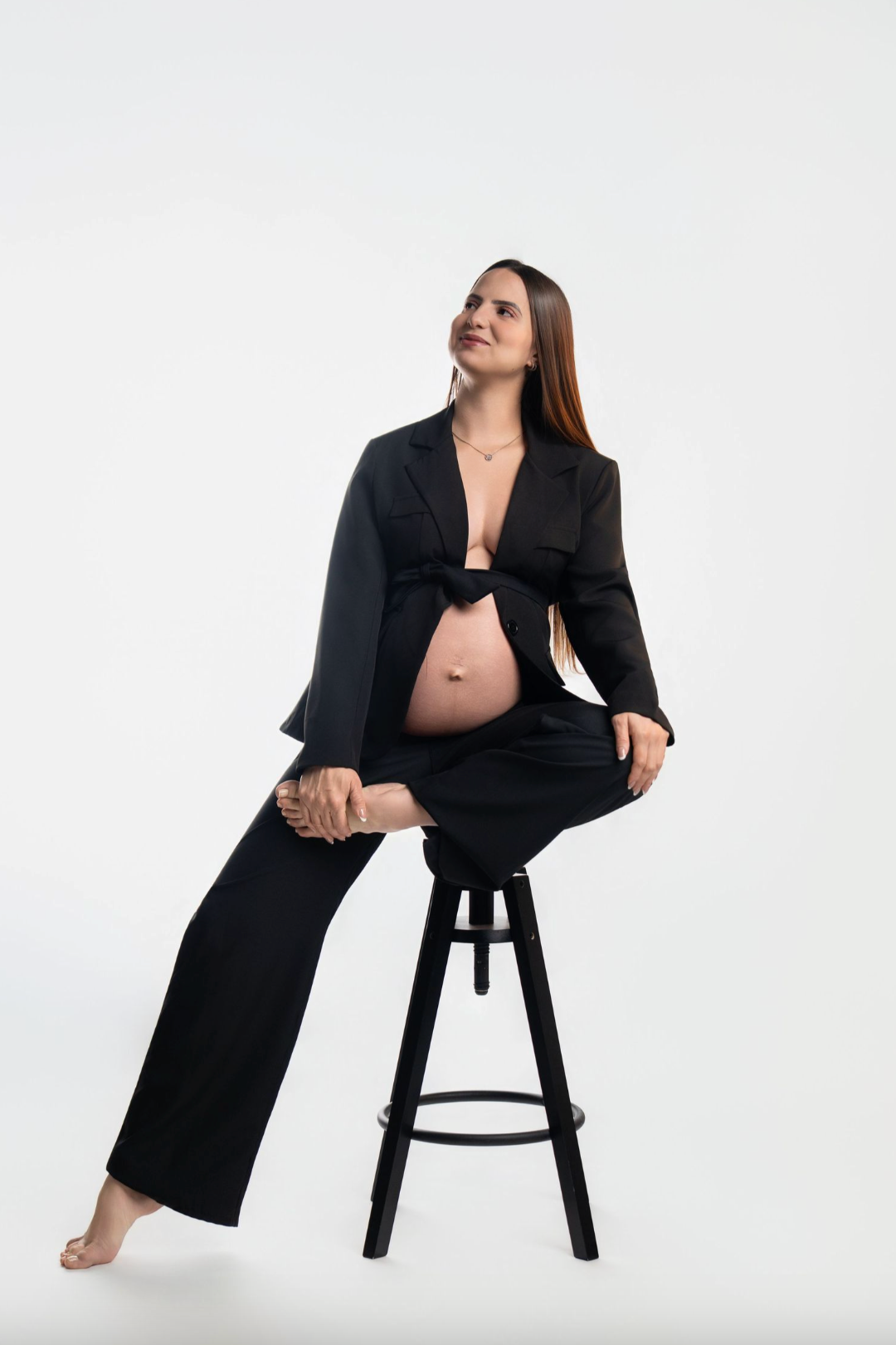 Pregnant woman in black blazer and pants sitting on stool, showing bare belly, against white background.