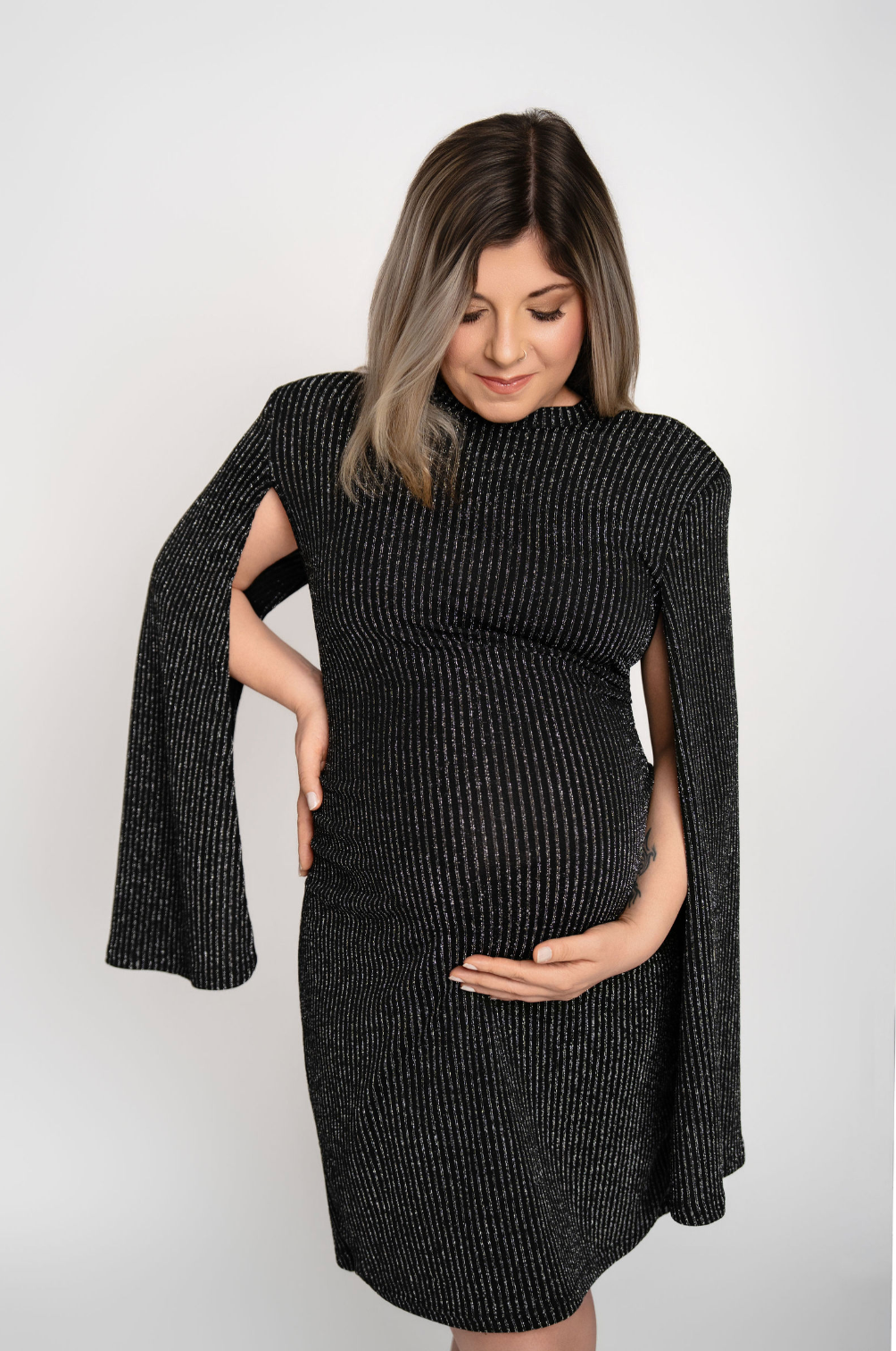 Pregnant woman in black shimmer cape dress cradling baby bump, maternity fashion photo on white background.